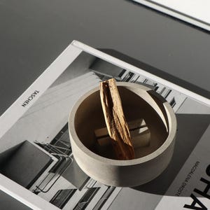 Könnte beinhalten: Ein Set aus drei Keramikbechern mit gesprenkelter, matter Oberfläche. Die Becher sind in Creme-, Hellgrau- und Dunkelgrautönen gehalten. Jeder Becher hat ein schlichtes, minimalistisches Design.