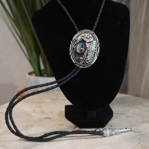 Op de afbeelding: Een bolo-das met een ovaal zilveren middendeel met reliëf van bloemen- en bladpatronen. Het midden bevat een kleine inleg van turkoois, koraal en onyx. Het koord is gemaakt van gevlochten zwart leer met zilveren metalen uiteinden.