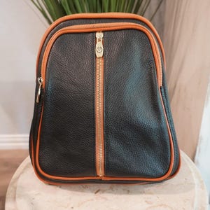 May include: A black backpack with a textured surface and tan piping trim. The bag features a central vertical gold-toned zipper with a circular logo pull, two main compartments with gold-toned zippers, and a rounded top silhouette.