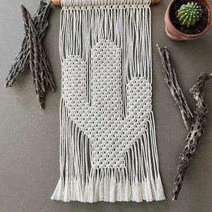 May include: A white macrame wall hanging in the shape of a cactus with a wooden dowel at the top. The hanging has a fringe at the bottom.
