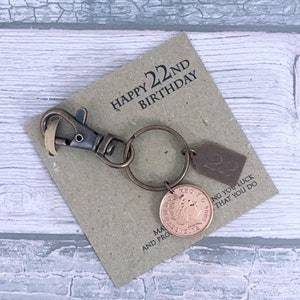 Könnte beinhalten: Eine braune Karte mit dem Text "Happy 22nd Birthday" und ein kleines braunes Leder-Etikett mit dem Text "Good luck that you do", das an einem Schlüsselring mit einer kupferfarbenen Münze befestigt ist.