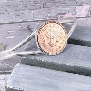 May include: A silver bracelet with a copper-coloured coin charm featuring a portrait of Queen Elizabeth II. The coin is dated 1963 and reads "ELIZABETH II D.G. REG. F.D." around the edge.