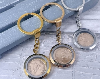 Puede incluir: Llaveros con colgantes de monedas. Los llaveros tienen un acabado dorado o plateado, con un colgante redondo que contiene una moneda. Las monedas muestran un diseño de pájaro y los años 1944 o 1947. Sobre una superficie azul claro.