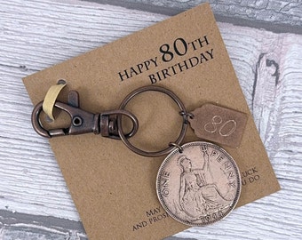 Puede incluir: Una tarjeta marrón con el texto "Happy 80th Birthday" y un llavero con un anillo de llave de color cobre, una etiqueta de color cobre con el número "80" en ella y una moneda de cobre con el texto "One Penny" y el año "1944".
