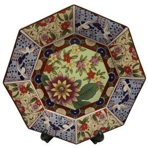 Puede incluir: Un plato decorativo octogonal con un diseño floral y de pájaros. El plato presenta un motivo floral central en tonos morados, amarillos y verdes, rodeado de paneles con flores y pájaros. El plato está sostenido por un soporte negro.