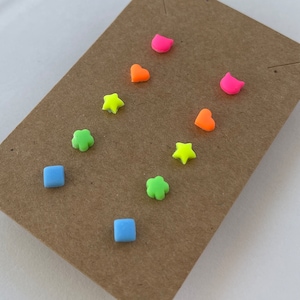 May include: A set of eight colorful earrings in various shapes, including stars, hearts, flowers, and squares. The earrings are made of a bright neon material and are displayed on a brown card.