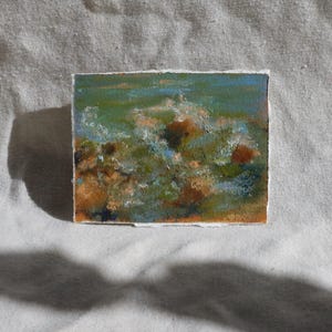 May include: A small, square painting with a textured surface. The artwork features a landscape scene with shades of green, blue, and brown. The painting is set against a white background.