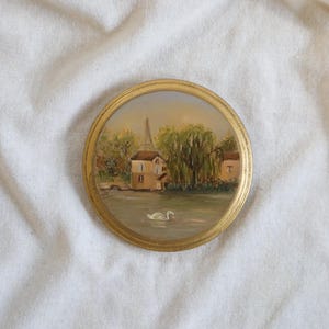 May include: A round, vintage-style painting with a gold frame. The artwork depicts a serene waterscape with buildings, trees, and two white swans. The painting has a soft, impressionistic style, with muted colors and a focus on light and shadow.