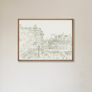 May include: A framed print featuring a sketch of a European cityscape. The artwork, rendered in shades of green, depicts buildings, trees, and a street lamp. The print is encased in a brown wooden frame, adding a touch of warmth to the piece.