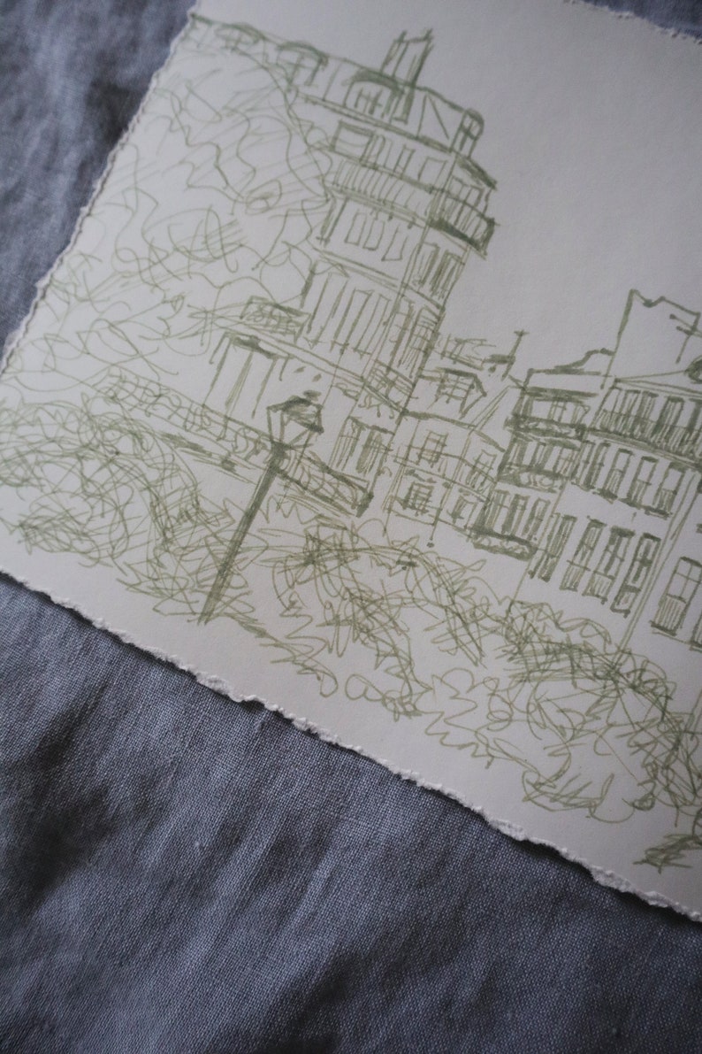 May include: A sketch of a European city scene, featuring buildings with balconies and a street lamp. The artwork is rendered in green ink on a textured, off-white paper with a torn edge. The drawing style is loose and impressionistic.