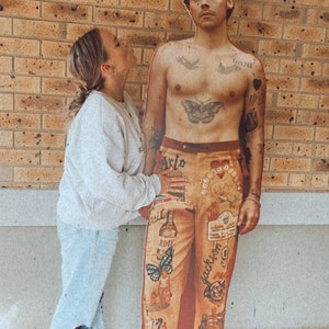 May include: A person is standing next to a cardboard cutout of a man with tattoos and colorful pants. The cutout has the words "Arlo Parks" and "ODE" on the pants.