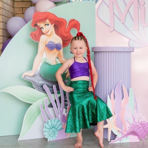 Op de afbeelding: Een jong meisje verkleed als zeemeermin met een groene staart en een paarse top. Ze staat voor een achtergrond met een cartoonafbeelding van Ariel uit De Kleine Zeemeermin.