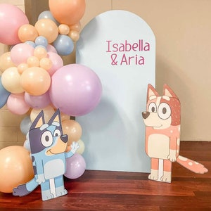 K&ouml;nnte beinhalten: Ein blauer und schwarzer Cartoon-Hund und ein brauner und wei&szlig;er Cartoon-Hund stehen vor einem hellblauen Hintergrund mit dem Text "Isabella & Aria" in Pink. Der Hintergrund ist mit einem Ballonbogen aus pastellrosa, pfirsichfarbenen und blauen Ballons geschm&uuml;ckt.