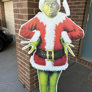 May include: A green Grinch character dressed as Santa Claus with a red suit, white fur trim, and a black belt. The Grinch is standing in front of a brick wall.