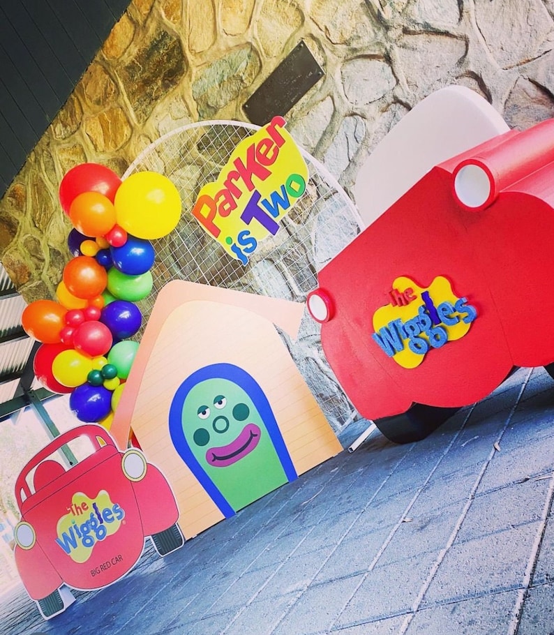 K&ouml;nnte beinhalten: Ein Geburtstags-Party-Setup mit einem roten und gelben 'The Wiggles'-Auto, einem roten und wei&szlig;en 'The Wiggles'-Schild, einem gelben und roten 'Parker is Two'-Schild und einem wei&szlig;en Ballonbogen mit roten, gelben, orangen und gr&uuml;nen Ballons.