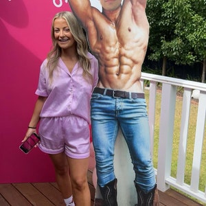 May include: A woman in a lavender satin pajama set stands next to a cardboard cutout of a shirtless man wearing a cowboy hat and jeans. The cutout is holding a bottle of beer. The woman is wearing white shoes with pink cowboy boot designs. The background is a pink wall with the words "Addis' Last Rodeo" written on it.