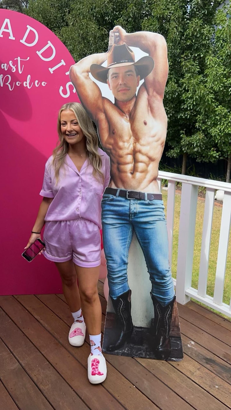 May include: A woman in a lavender satin pajama set stands next to a cardboard cutout of a shirtless man wearing a cowboy hat and jeans. The cutout is holding a bottle of beer. The woman is wearing white shoes with pink cowboy boot designs. The background is a pink wall with the words "Addis' Last Rodeo" written on it.
