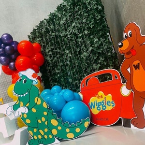 K&ouml;nnte beinhalten: Ein gr&uuml;ner Dinosaurier mit gelben Flecken, ein rotes Auto mit den Worten "The Wiggles Big Red Car" und ein brauner Hund mit roter Zunge und dem Buchstaben "W" auf der Brust. Der Hintergrund ist eine gr&uuml;ne Efeuwand und es gibt blaue und rote Ballons.