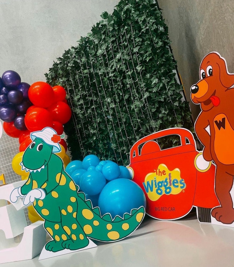 K&ouml;nnte beinhalten: Ein gr&uuml;ner Dinosaurier mit gelben Flecken, ein rotes Auto mit den Worten "The Wiggles Big Red Car" und ein brauner Hund mit roter Zunge und dem Buchstaben "W" auf der Brust. Der Hintergrund ist eine gr&uuml;ne Efeuwand und es gibt blaue und rote Ballons.