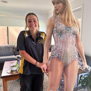 May include: A young person wearing a black and yellow uniform shirt and black pants is standing next to a cardboard cutout of a pop star wearing a sparkly silver and pink bodysuit. The pop star is holding a microphone.