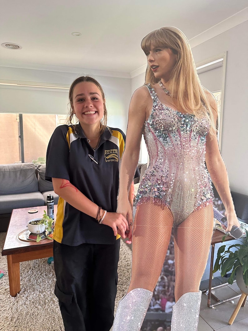 May include: A young person wearing a black and yellow uniform shirt and black pants is standing next to a cardboard cutout of a pop star wearing a sparkly silver and pink bodysuit. The pop star is holding a microphone.
