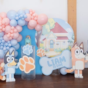 K&ouml;nnte beinhalten: Ein blauer und rosa Ballonbogen mit einem blauen und wei&szlig;en knochenf&ouml;rmigen Schild, auf dem "Liam ist eins" steht. Es gibt zwei Comic-Hundfiguren, eine blaue und eine orange, und eine blaue und wei&szlig;e Hauskulisse.