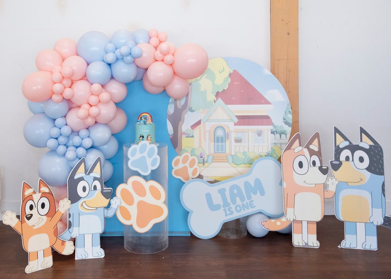 K&ouml;nnte beinhalten: Ein blauer und rosa Ballonbogen mit einem blauen und wei&szlig;en knochenf&ouml;rmigen Schild, auf dem "Liam ist eins" steht. Es gibt zwei Comic-Hundfiguren, eine blaue und eine orange, und eine blaue und wei&szlig;e Hauskulisse.