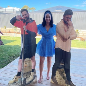 May include: Three cardboard cutouts of people standing on a wooden deck. The person in the middle is a woman wearing a blue dress with white polka dots. The person on the left is a man wearing a black shirt, orange vest, and brown boots. The person on the right is a man wearing a beige shirt, black pants, and brown boots.