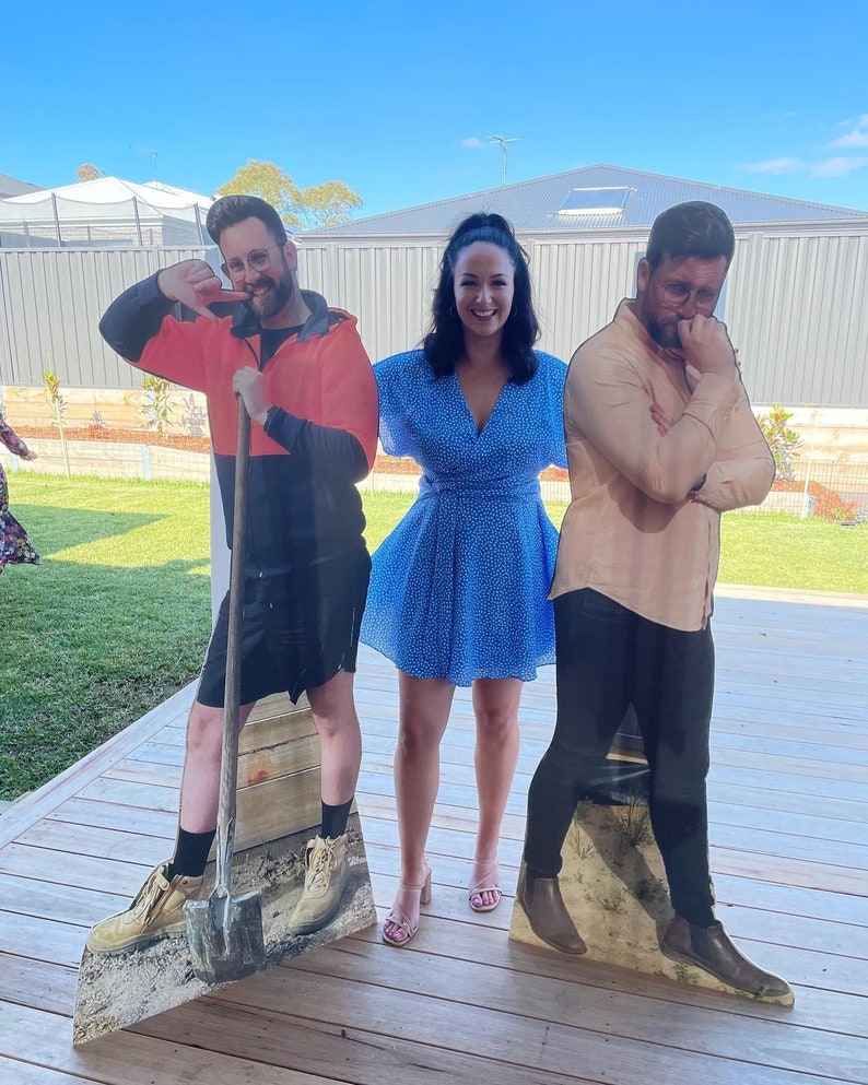 May include: Three cardboard cutouts of people standing on a wooden deck. The person in the middle is a woman wearing a blue dress with white polka dots. The person on the left is a man wearing a black shirt, orange vest, and brown boots. The person on the right is a man wearing a beige shirt, black pants, and brown boots.