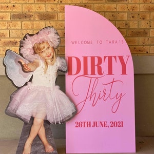 May include: Pink and red party sign with the text "Welcome to Tara's Dirty Thirty 26th June, 2021". A cutout of a young girl dressed as a fairy is standing next to the sign.