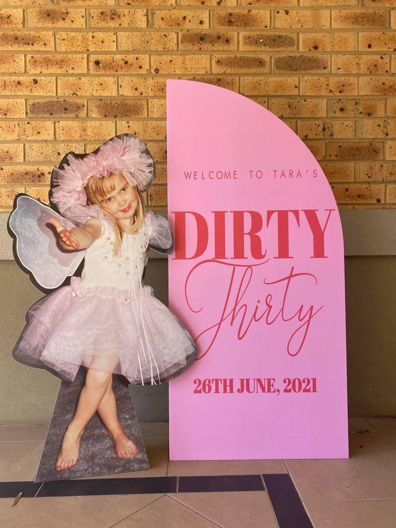 May include: Pink and red party sign with the text "Welcome to Tara's Dirty Thirty 26th June, 2021". A cutout of a young girl dressed as a fairy is standing next to the sign.