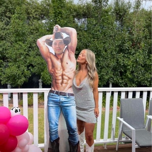 May include: A woman in a sparkly silver dress and white cowboy boots poses with a cardboard cutout of a shirtless man wearing jeans and a cowboy hat.