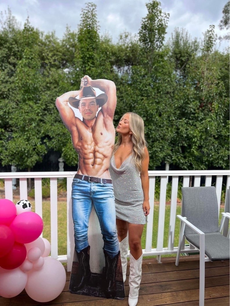 May include: A woman in a sparkly silver dress and white cowboy boots poses with a cardboard cutout of a shirtless man wearing jeans and a cowboy hat.