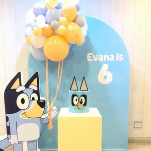 K&ouml;nnte beinhalten: Ein blauer und gelber Geburtstagsparty-Hintergrund mit einem blauen und wei&szlig;en Ballonbogen, einer blauen und gelben Torte mit einem Cartoon-Hundegesicht und einem Schild mit der Aufschrift "Evana Is 6".
