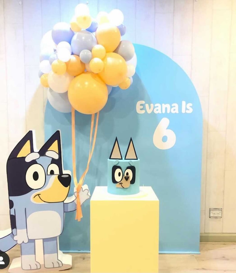 K&ouml;nnte beinhalten: Ein blauer und gelber Geburtstagsparty-Hintergrund mit einem blauen und wei&szlig;en Ballonbogen, einer blauen und gelben Torte mit einem Cartoon-Hundegesicht und einem Schild mit der Aufschrift "Evana Is 6".