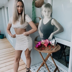 May include: A woman in a sparkly silver crop top and matching skirt poses in front of a cutout of a young girl. The woman is wearing black heels and has her hair down. There is a small table with a glass of wine and heart-shaped balloons in the background.