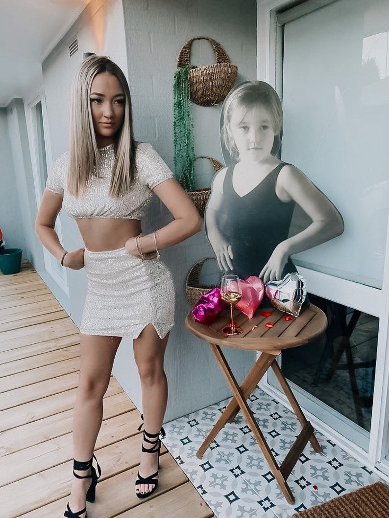 May include: A woman in a sparkly silver crop top and matching skirt poses in front of a cutout of a young girl. The woman is wearing black heels and has her hair down. There is a small table with a glass of wine and heart-shaped balloons in the background.
