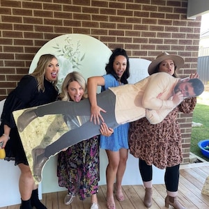May include: Four people are holding a cardboard cutout of a man. The cutout is wearing a light brown shirt and has a mustache. The people are all smiling and laughing. The cutout is being held up in front of a brick wall.