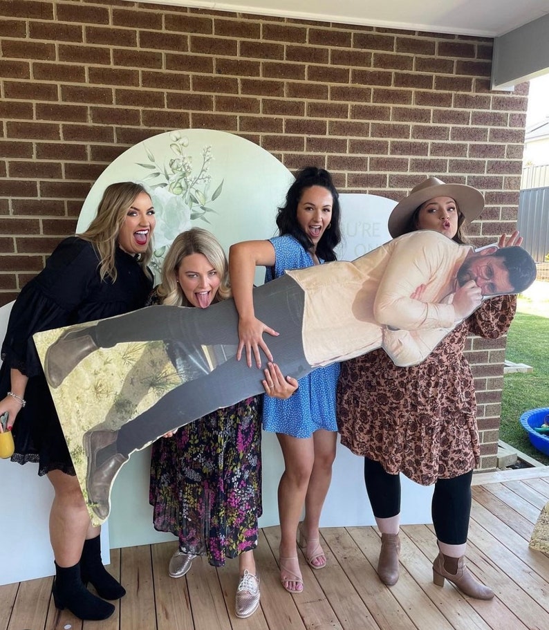 May include: Four people are holding a cardboard cutout of a man. The cutout is wearing a light brown shirt and has a mustache. The people are all smiling and laughing. The cutout is being held up in front of a brick wall.