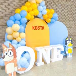 K&ouml;nnte beinhalten: Eine Geburtstagsparty-Kulisse mit einem gelben und blauen Ballonbogen, zwei gro&szlig;en Pappfiguren von Cartoon-Hunden und wei&szlig;en Schaumstoffbuchstaben, die "ONE" buchstabieren. Die Kulisse zeigt einen gelben und blauen Halbkreis mit dem Namen "KOSTA" in roten Buchstaben.