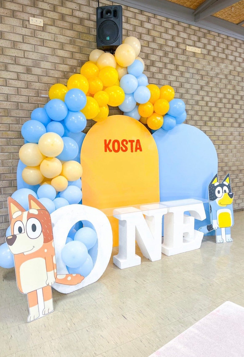 K&ouml;nnte beinhalten: Eine Geburtstagsparty-Kulisse mit einem gelben und blauen Ballonbogen, zwei gro&szlig;en Pappfiguren von Cartoon-Hunden und wei&szlig;en Schaumstoffbuchstaben, die "ONE" buchstabieren. Die Kulisse zeigt einen gelben und blauen Halbkreis mit dem Namen "KOSTA" in roten Buchstaben.