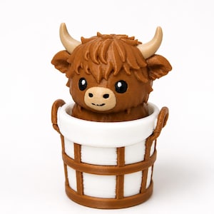 May include: A small 3D printed figurine of a brown Highland cow with curved tan horns, black eyes, and a light tan muzzle, sitting inside a white and brown bucket with two side handles. The cow has textured fur details and a small smile.