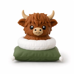 May include: A brown Highland cow figurine with shaggy fur, curved beige horns, and large black eyes. The cow is positioned inside a white circular ring, which rests on top of a square olive green cushion featuring small white corner tassels.