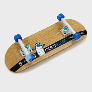 Puede incluir: Un monopatín de madera en miniatura con ruedas azules y ejes blancos. La tabla presenta un patrón de vetas de madera clara, una franja negra con el texto "COMPREADY" y un dibujo de yeti.