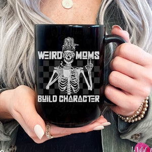 May include: Black ceramic coffee mug with a white graphic of a skeleton wearing sunglasses and a messy bun. The text "Weird Moms Build Character" is printed on the mug.