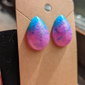 May include: A pair of teardrop-shaped earrings with a gradient of pink and blue glitter. The earrings are displayed on a brown card.