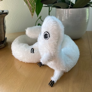 May include: A white plush toy shaped like a worm or snake, with black eyes and tiny black claws. The soft toy is on a light brown wooden surface, with green plants in the background.