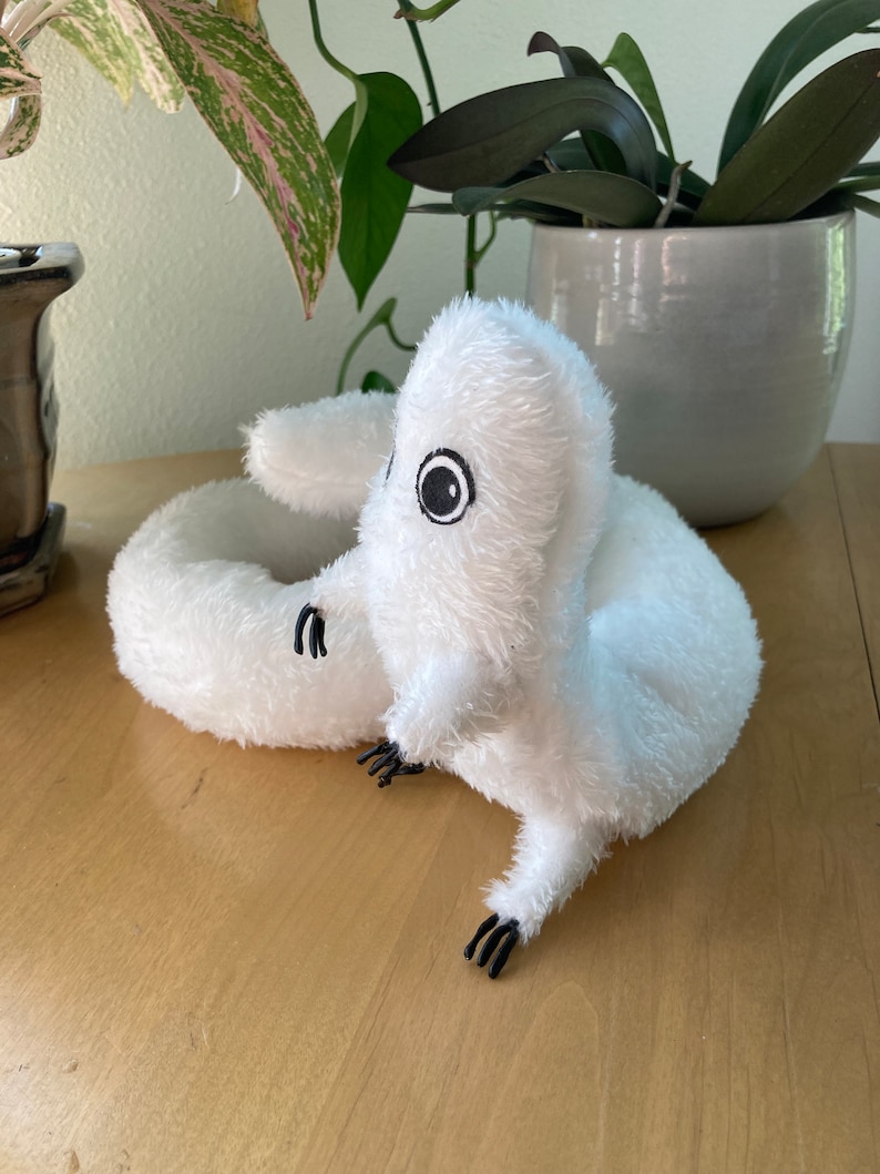 May include: A white plush toy shaped like a worm or snake, with black eyes and tiny black claws. The soft toy is on a light brown wooden surface, with green plants in the background.