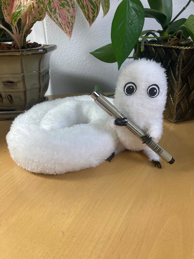 May include: A white plush creature with large black eyes and a pen held in its paws. The creature is lying on a wooden surface, with green plants in the background. The plush toy has a soft, furry texture.