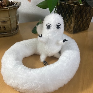 May include: A white plush toy shaped like a worm with a small, round body and a large, circular body. The toy has large, black eyes and small, black arms. The toy is sitting on a wooden surface, with plants in the background.